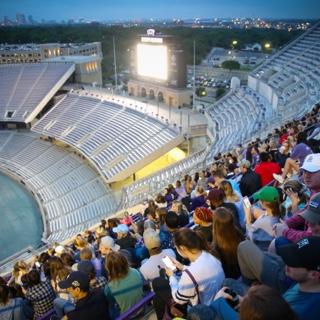 A group of graduating TCU seniors sits together in the high stadium bleachers as they watch the sun rise behind the lights of Fort Worth.